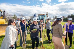 Several people holding shovels and wearing hardhats gather in a field with construction equipment.