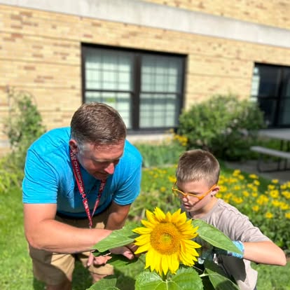 S young child examines a sunflower outside with a teacher.