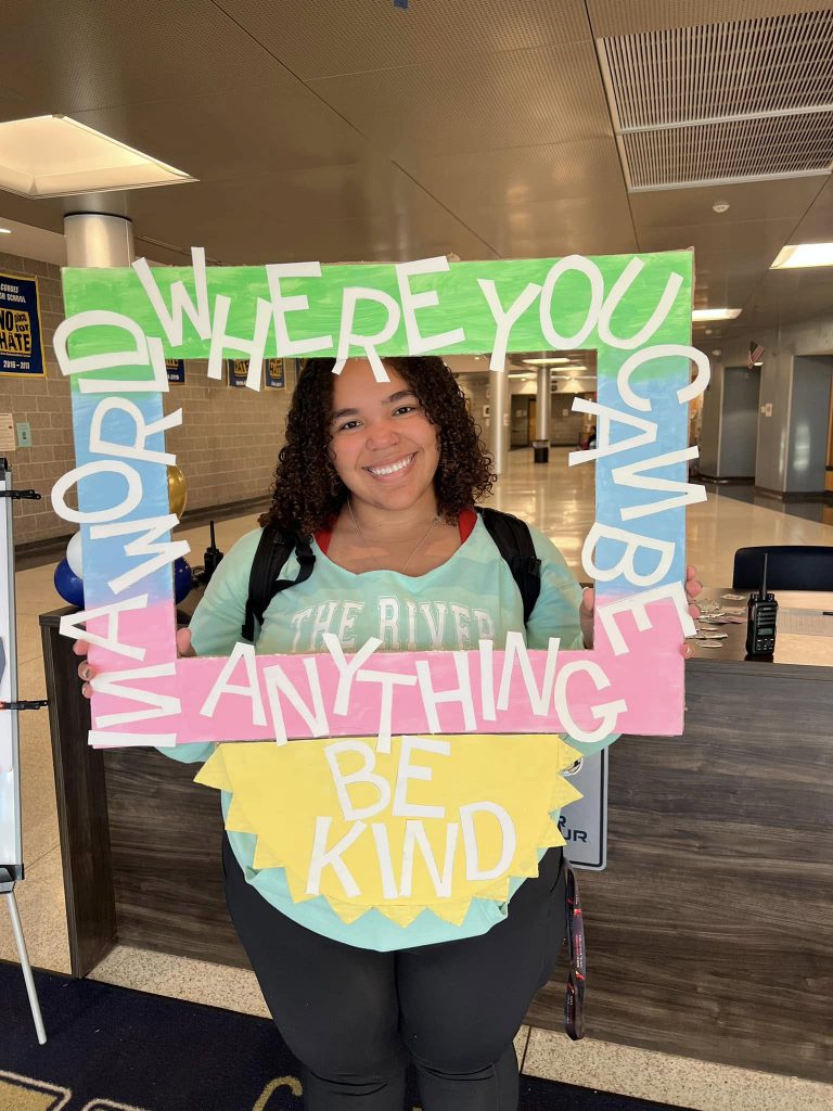 A student smiles inside a handheld frame that say "In a world where you can be anything be kind".