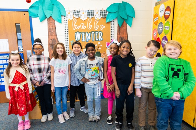 Elementary school children stand in front of a bulletin board smiling.