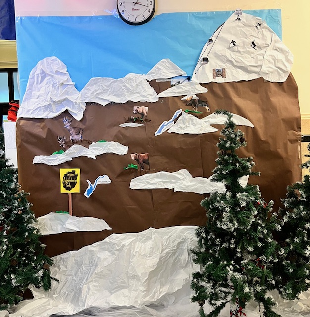 A craft paper mountain with cutout animals and skiers with some fake pine trees in front of it.